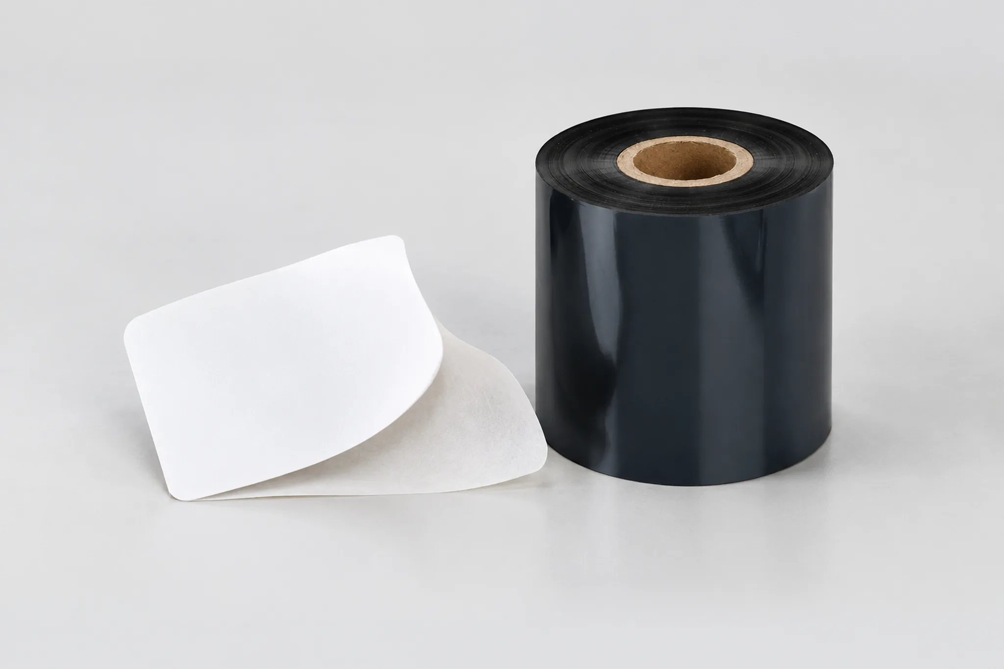 Close-up of a black ribbon roll beside a blank adhesive label partly lifted from its release liner.
