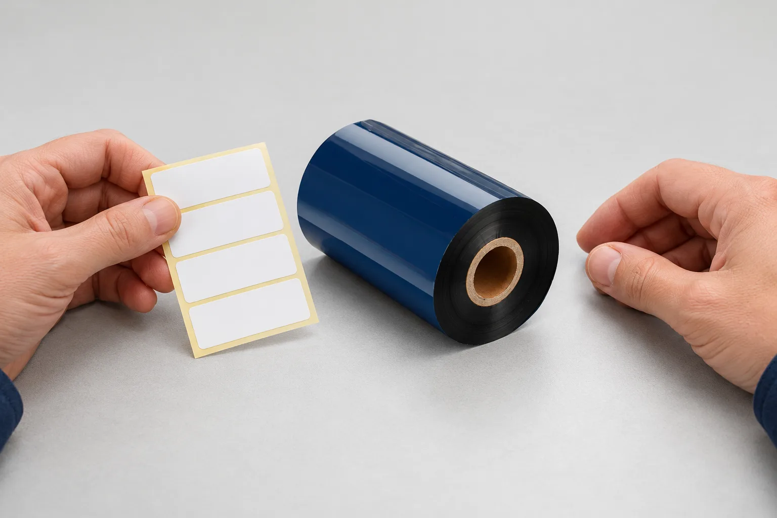 Hands comparing a blank label sheet with a separate black thermal transfer ribbon roll at a workbench.